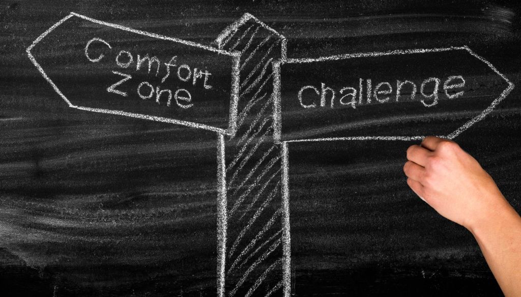 Chalkboard drawing of a hand choosing between two arrows labeled comfort zone and challenge, symbolizing embracing challenges over staying comfortable.