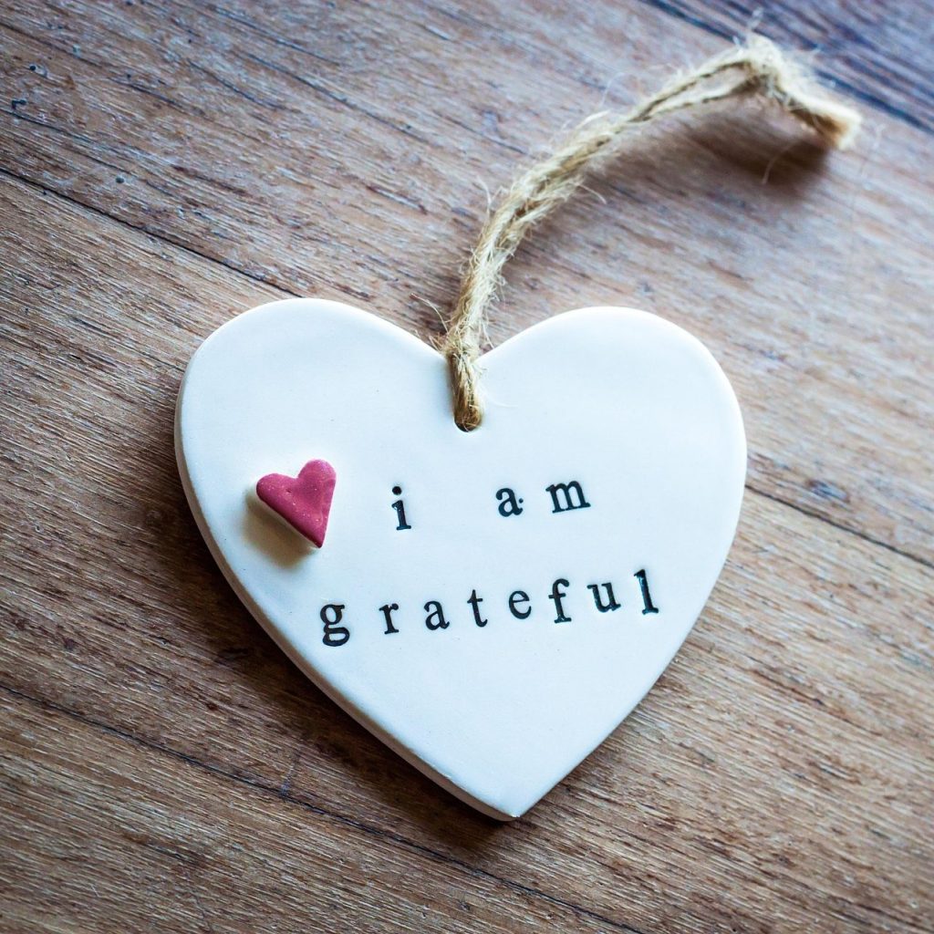 White ceramic heart ornament with a small red heart and the words “i am grateful” stamped on it, resting on a wooden surface—symbolizing gratitude, mindfulness, and positive affirmations.